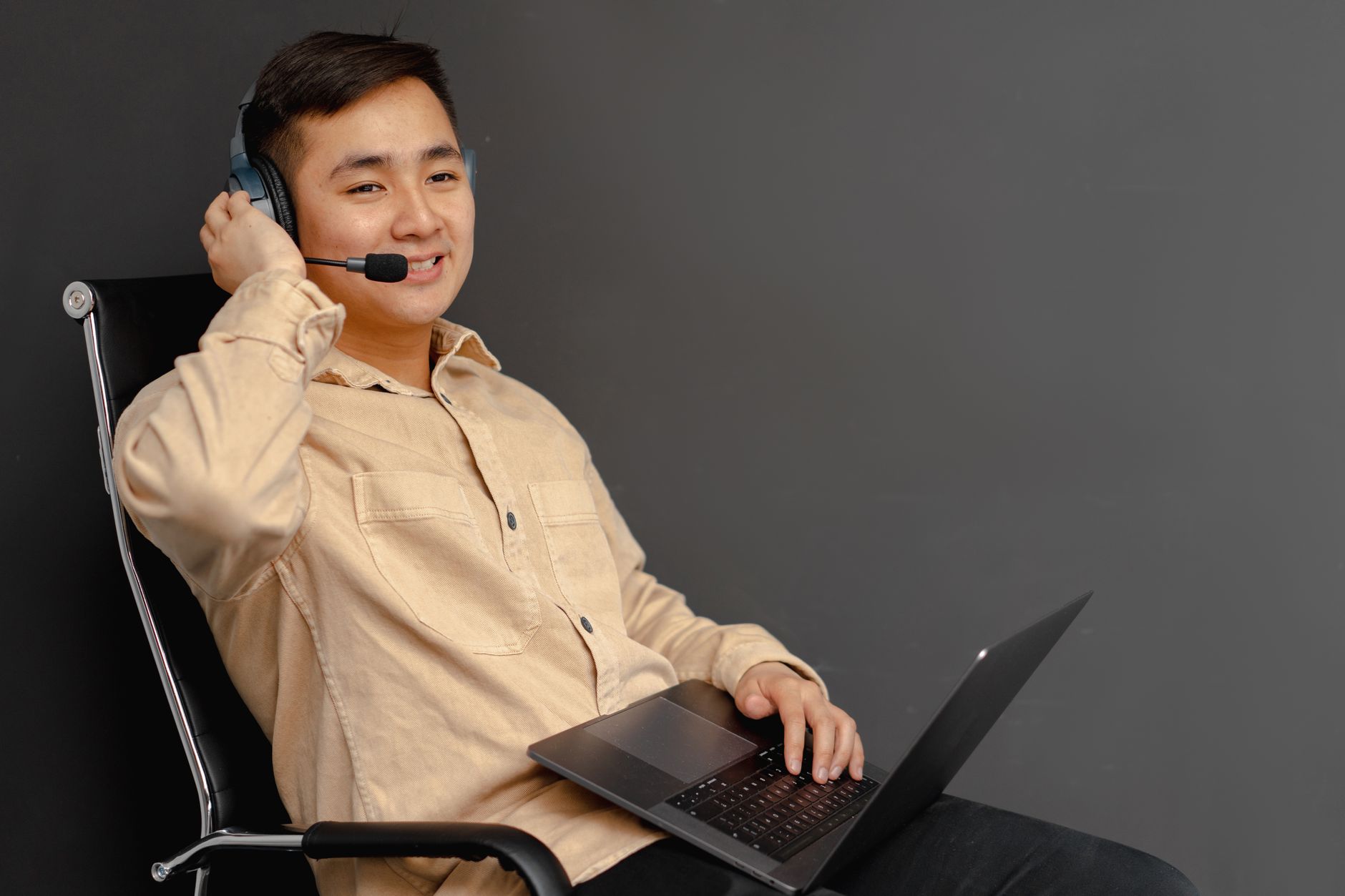 Joven sonriente con auriculares y micrófono, sentado en una silla con un portátil sobre las rodillas