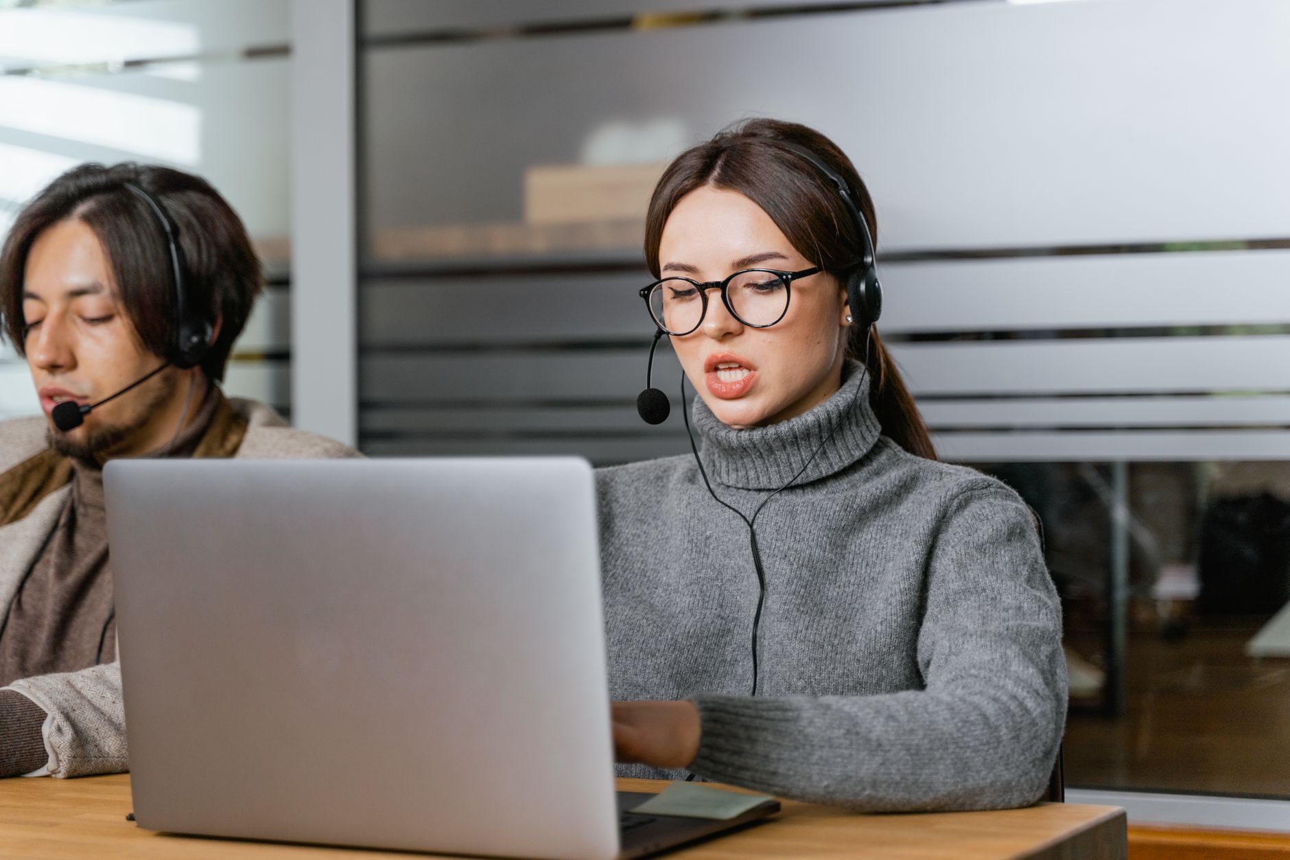 Mujer con gafas y auriculares con micrófono hablando mientras trabaja frente a su portátil